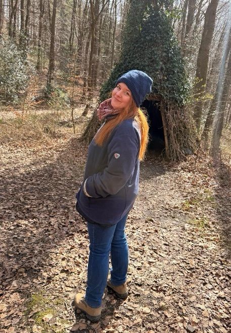Eine Person mit langen roten Haaren, die eine blaue Mütze und Jacke trägt, steht in einem sonnigen, laubbedeckten Wald.