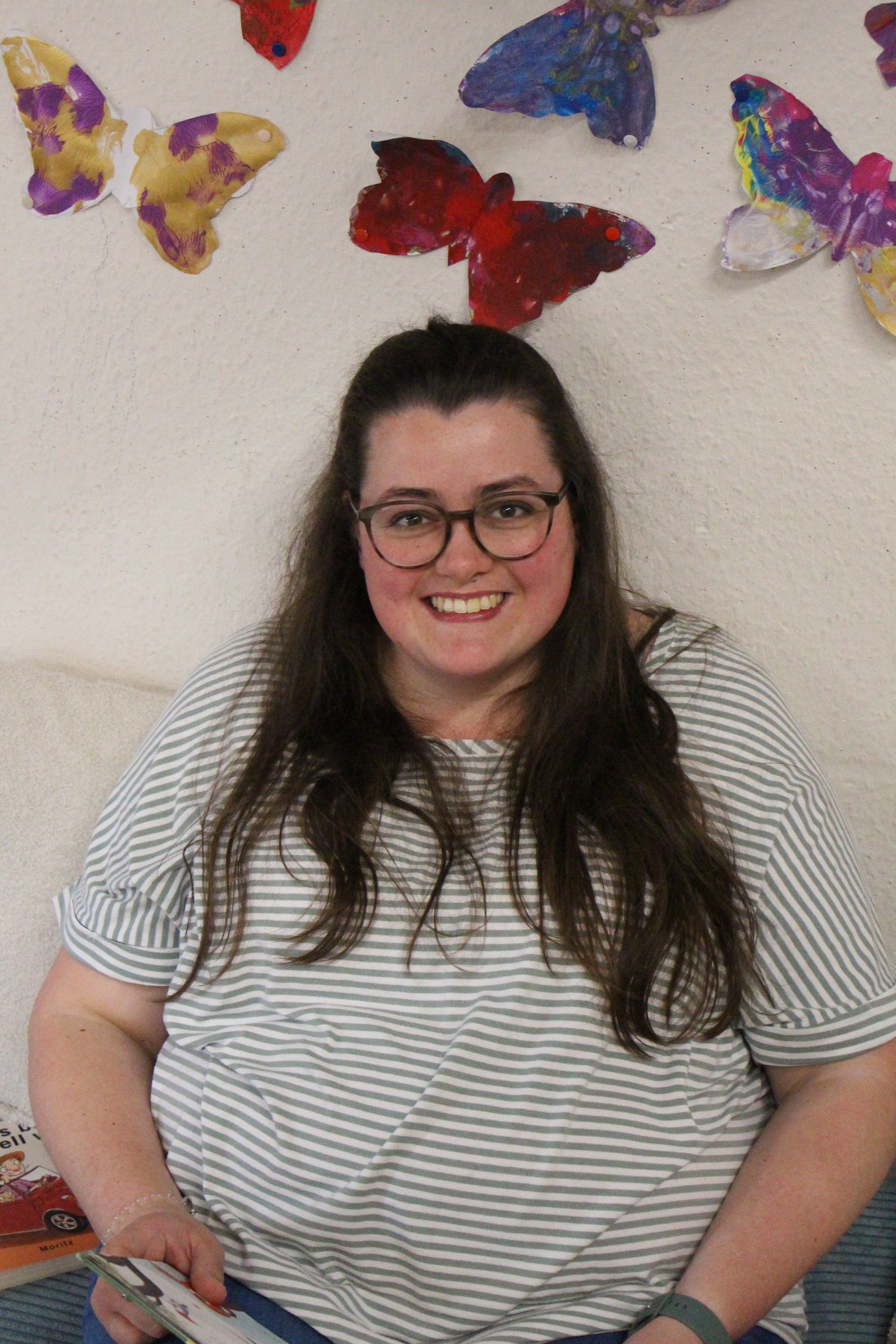 Eine lächelnde Person mit langen dunklen Haaren und Brille sitzt vor einer weißen Wand, die mit Papierschmetterlingen verziert ist.