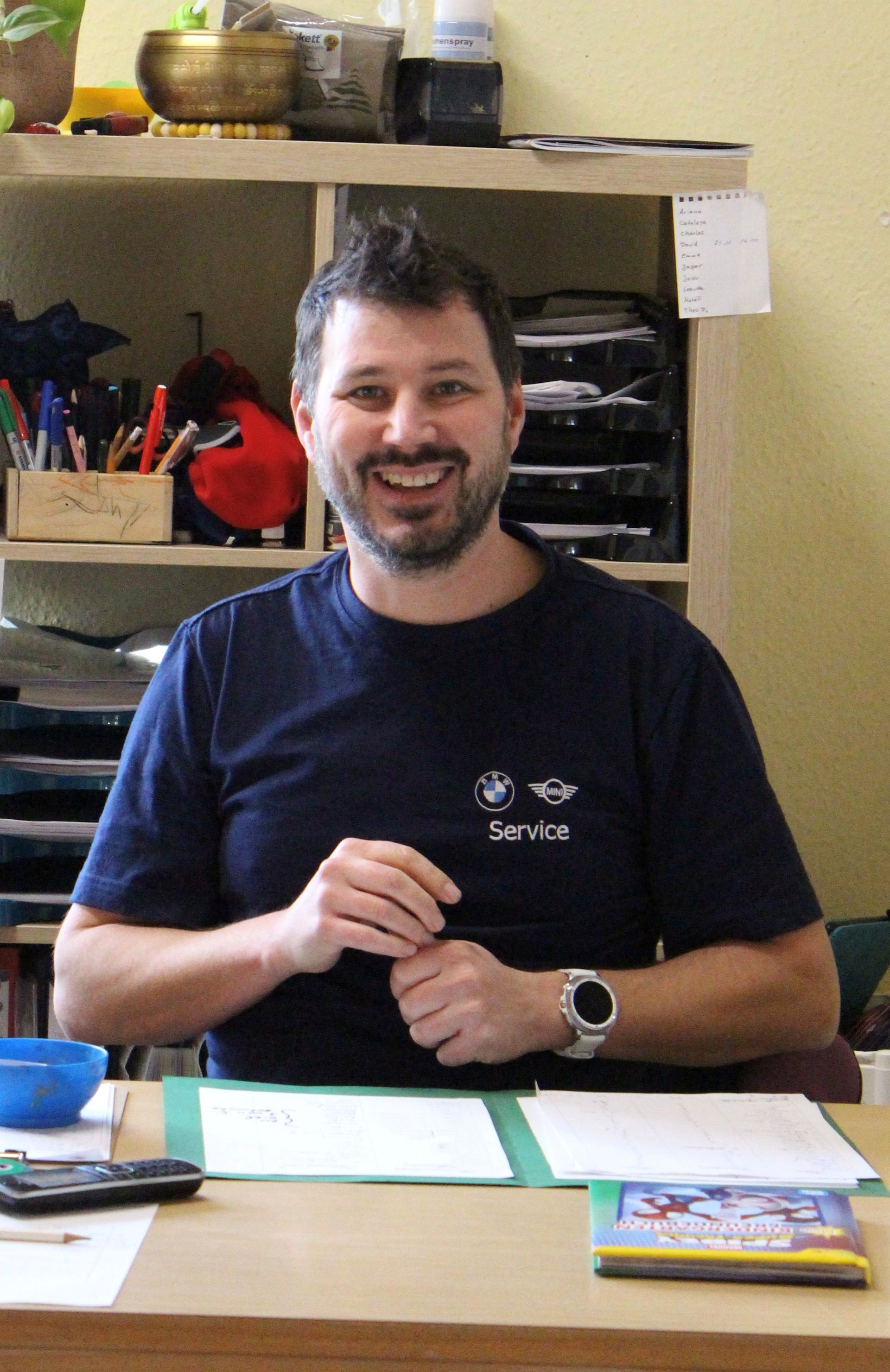 Eine lächelnde Person in einem dunkelblauen T-Shirt sitzt an einem Schreibtisch mit Papieren und Stiften in einem Gruppenraum des kindergartens.