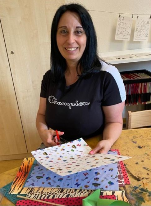 Eine Person lächelt, während sie an einem Tisch mit verschiedenen bunten Bastelmaterialien gemustertes Papier ausschneidet.