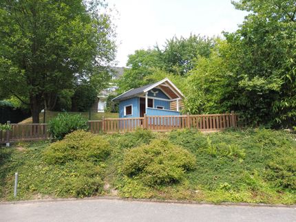 Ein kleines, blaues Holzspielhaus mit weiß umrandetem Dach steht auf einem grasbewachsenen Hang hinter einem Holzzaun.