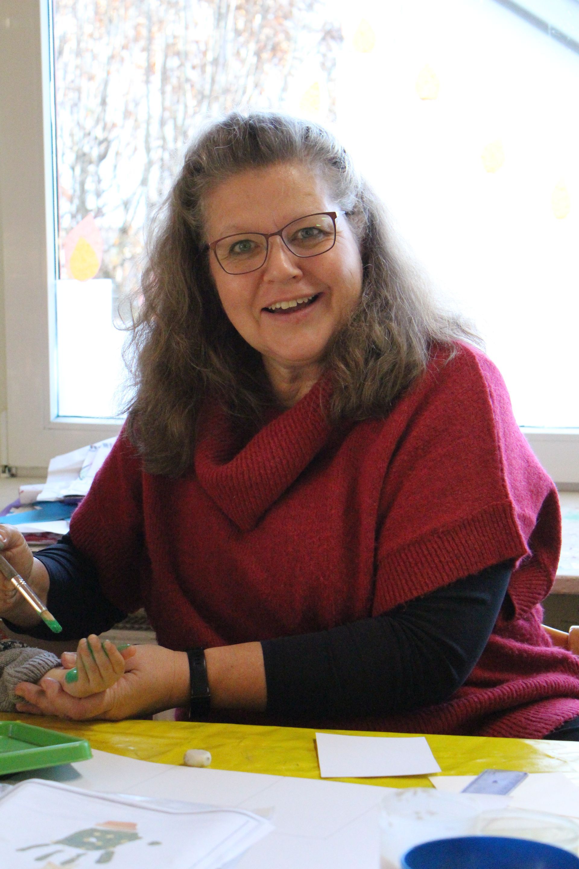 Eine lächelnde Person in einem roten Pullover bemalt eine Kinderhand, während sie an einem Tisch mit Malutensilien sitzt.