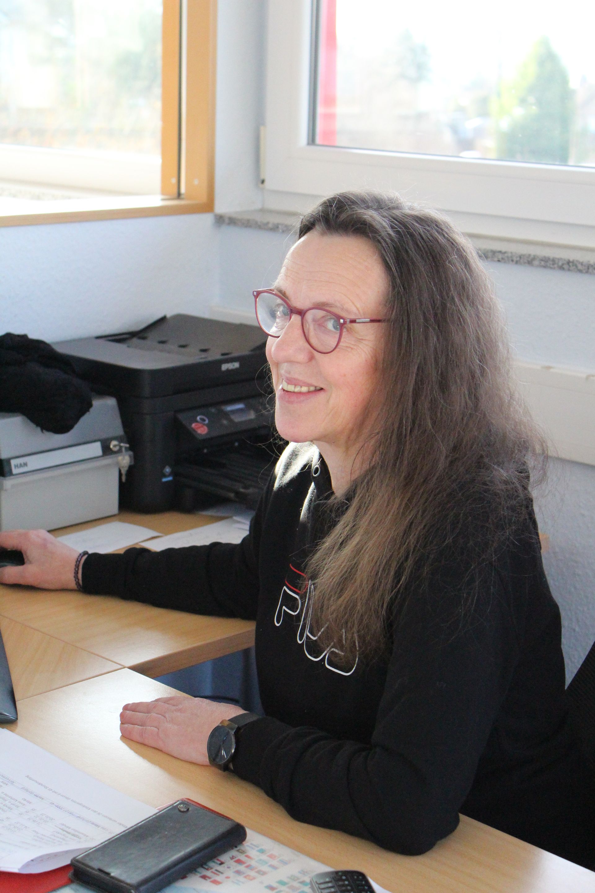 Eine lächelnde Person mit langen, gewellten Haaren trägt eine Brille und ein schwarzes Hemd und arbeitet an einem Schreibtisch in einem Büro.