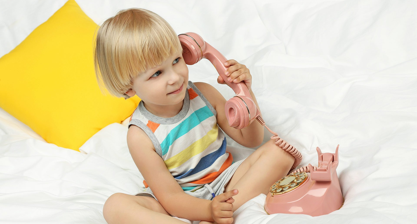 Eine Junge sitzt lächelnd auf weißer Bettwäsche und hält dabei ein altmodisches rosa Telefon in der Hand.