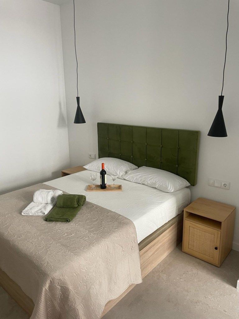 Dormitorio con cabecero verde oliva, dos mesitas de noche y una botella de vino sobre la cama. Dos lámparas colgantes negras cuelgan encima.