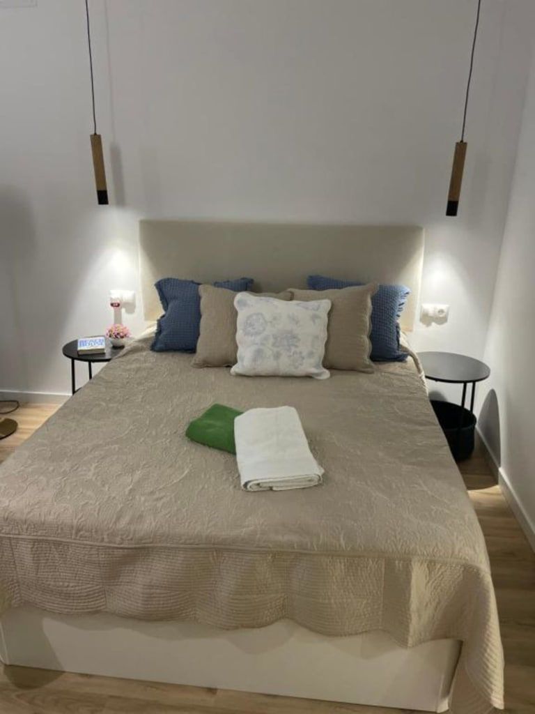 Dormitorio con cama hecha, edredón beige y varias almohadas. Dos mesitas de noche flanquean la cama y lámparas colgantes cuelgan encima.