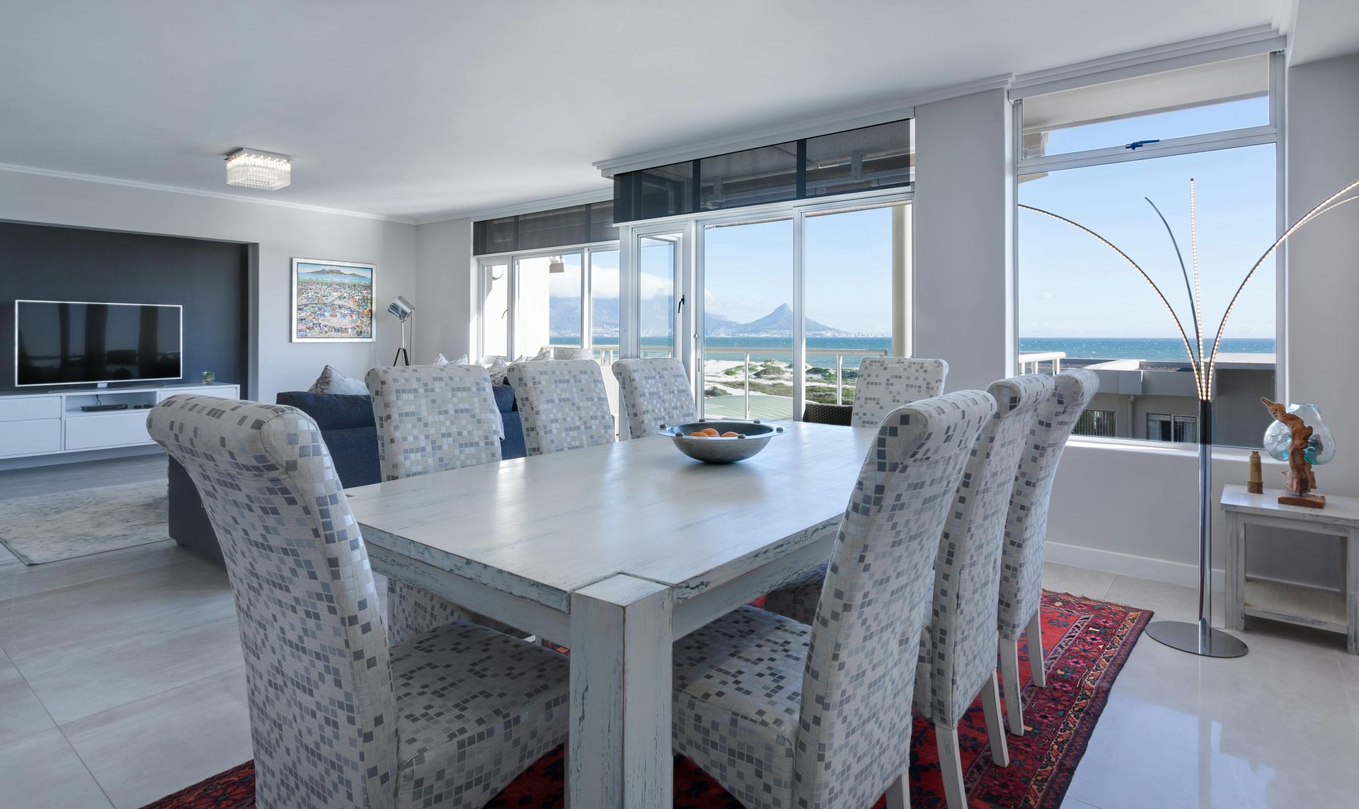 Comedor con una gran mesa blanca preparada para comer, con sillas estampadas, frente a una gran ventana con vista a un cuerpo de agua y montañas.