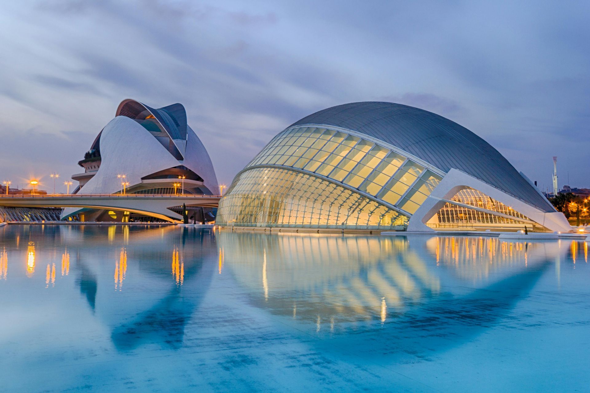 Arquitectura moderna reflejada en el agua al anochecer. Los edificios de Valencia, España, se iluminan contra un cielo azul oscuro.