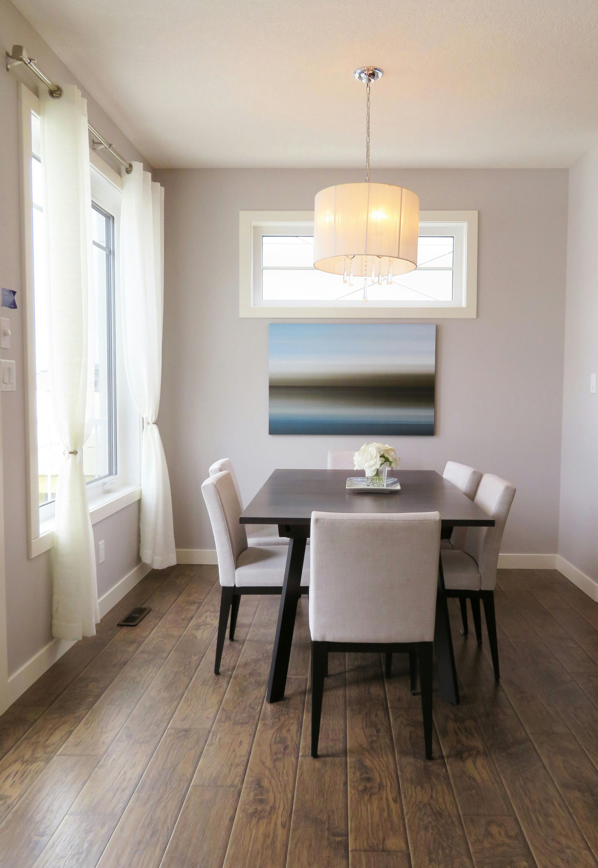 Comedor con mesa de madera oscura, sillas gris claro y lámpara, frente a una ventana y una obra de arte.