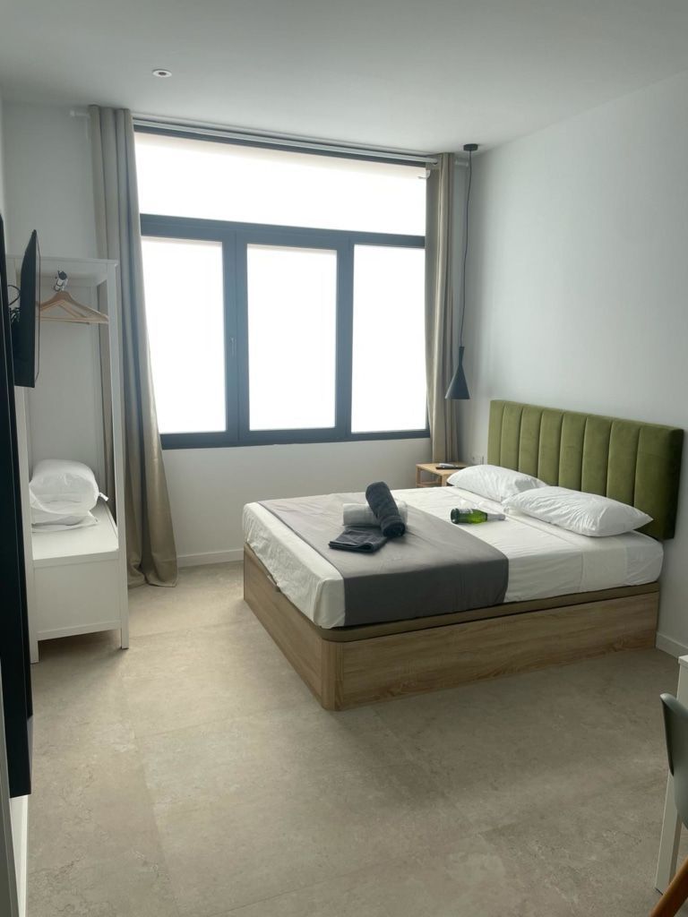 Un dormitorio con cama, ventana y mesita de noche. La cama tiene un cabecero verde y una base marrón claro, con toallas encima.