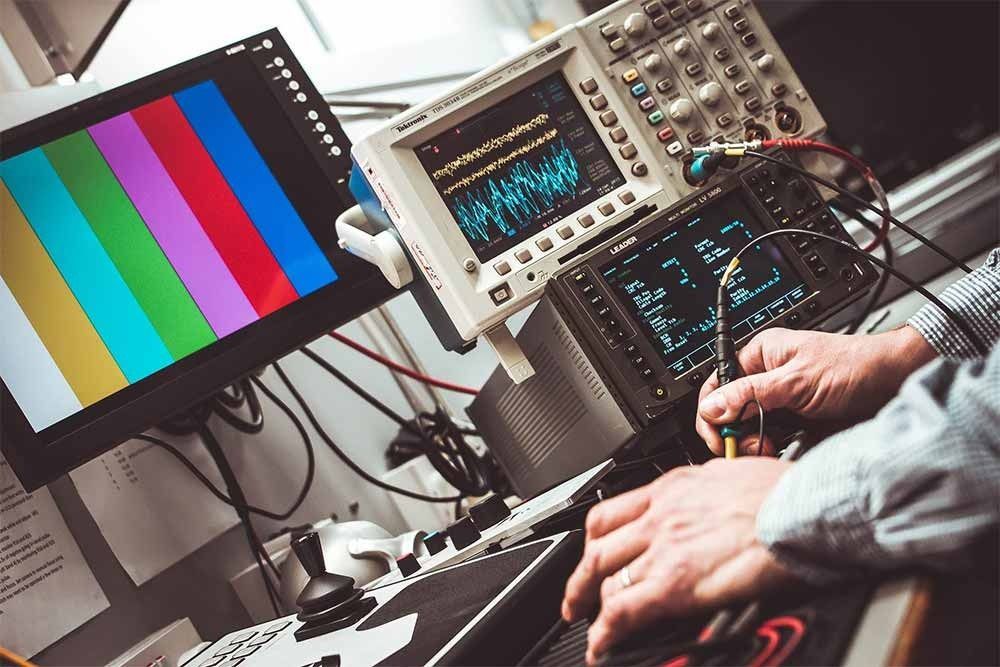 Una persona utiliza un destornillador en un laboratorio con equipos de prueba, un monitor que muestra barras de color y pantallas de osciloscopio.