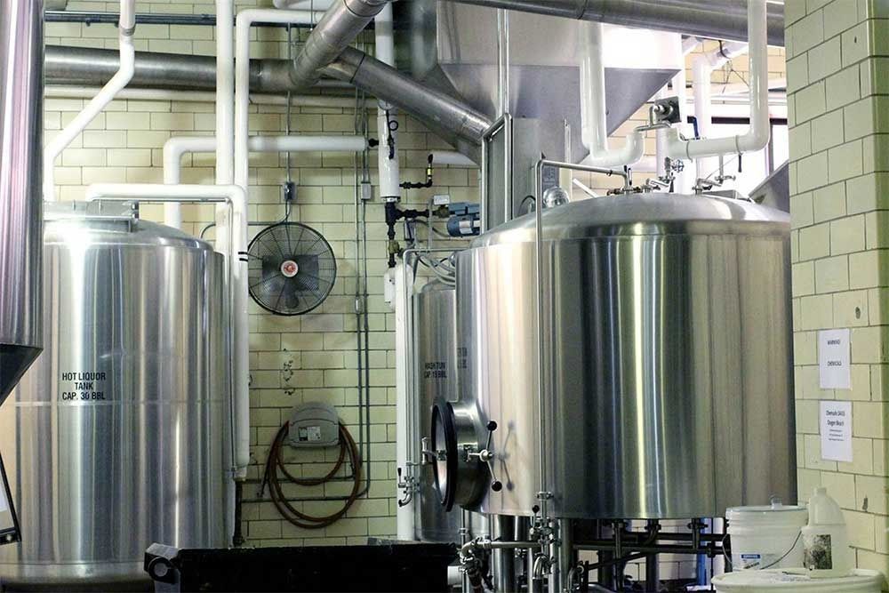 Tanques y tuberías de cervecería en un entorno industrial con un ventilador en la pared.