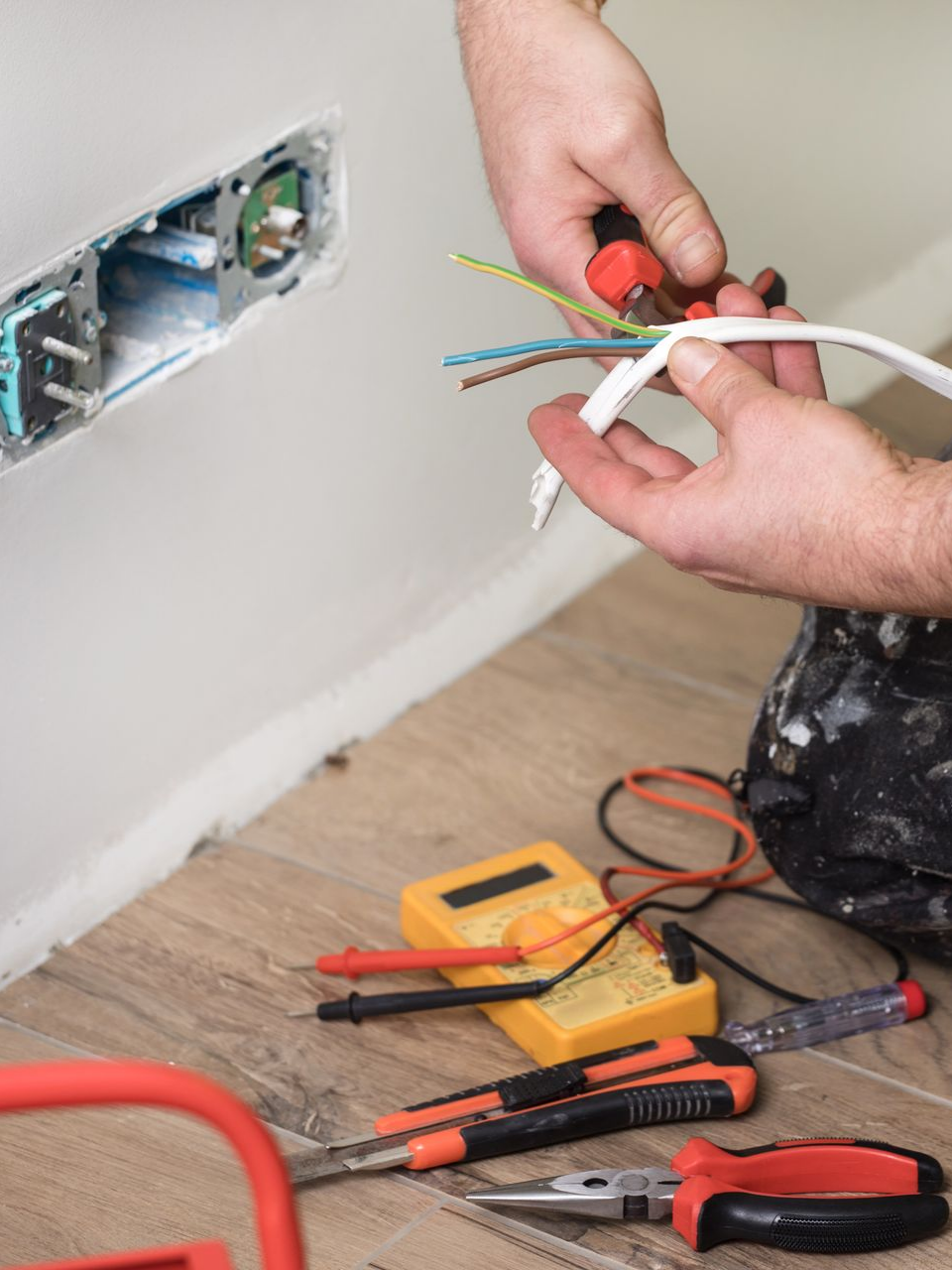 Un électricien installe une prise électrique ; mur blanc, sol en bois, outils à proximité, outils rouges et noirs, fils électriques.