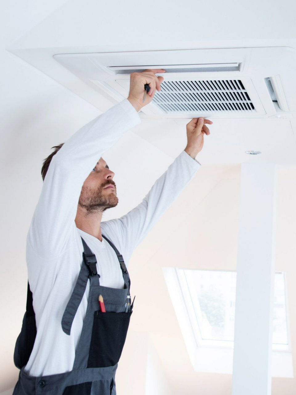 Un homme en salopette inspecte un climatiseur de plafond. Il tend les deux mains vers le haut.