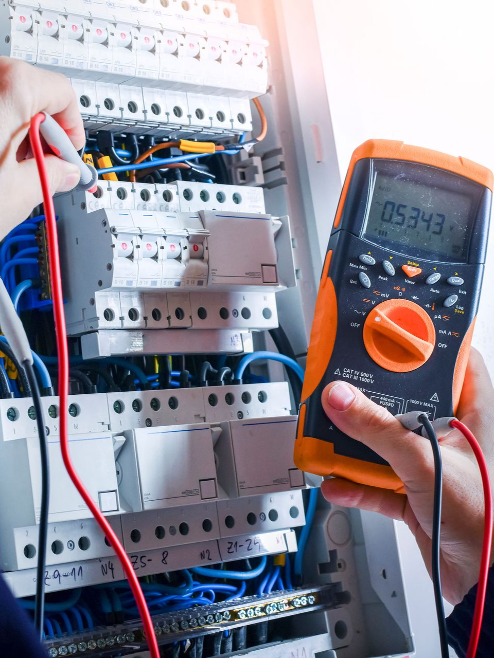 Une personne utilise un multimètre pour tester le câblage électrique d'un tableau électrique. Multimètre avec fils orange et noir, fils bleus et noirs.