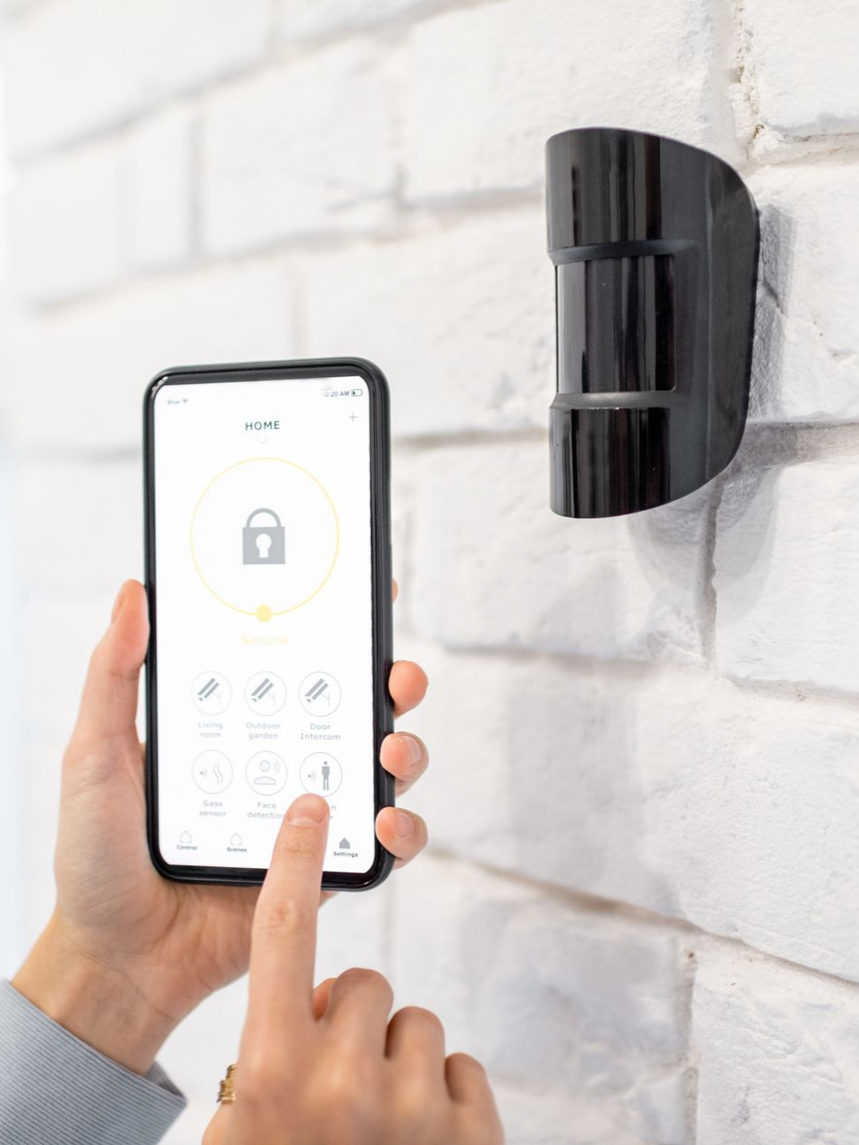 Main d'une personne utilisant une application pour smartphone afin de contrôler un capteur de sécurité fixé à un mur de briques blanches.