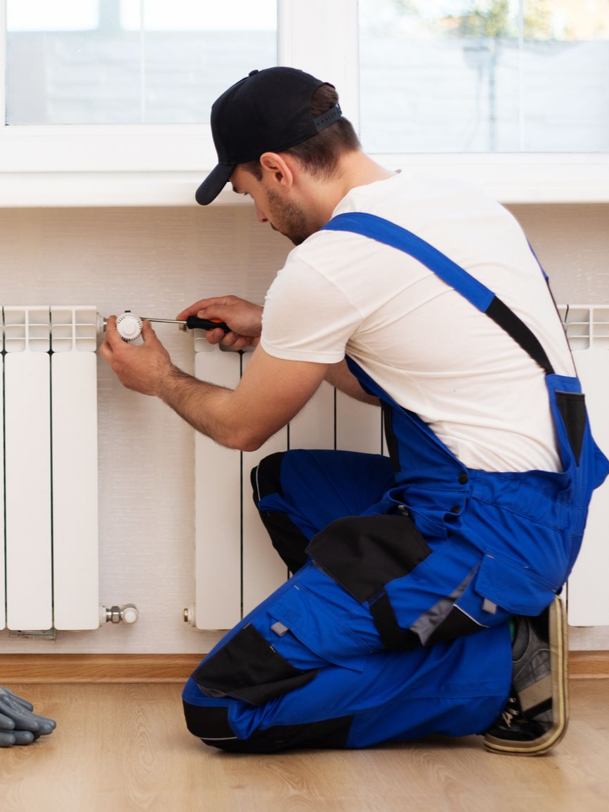 Plombier à genoux, réparant un radiateur blanc avec un tournevis. Il porte une combinaison bleue et une casquette noire.