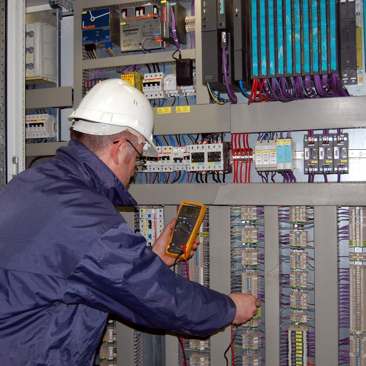 Un homme portant un casque de chantier utilise un multimètre sur un tableau électrique.