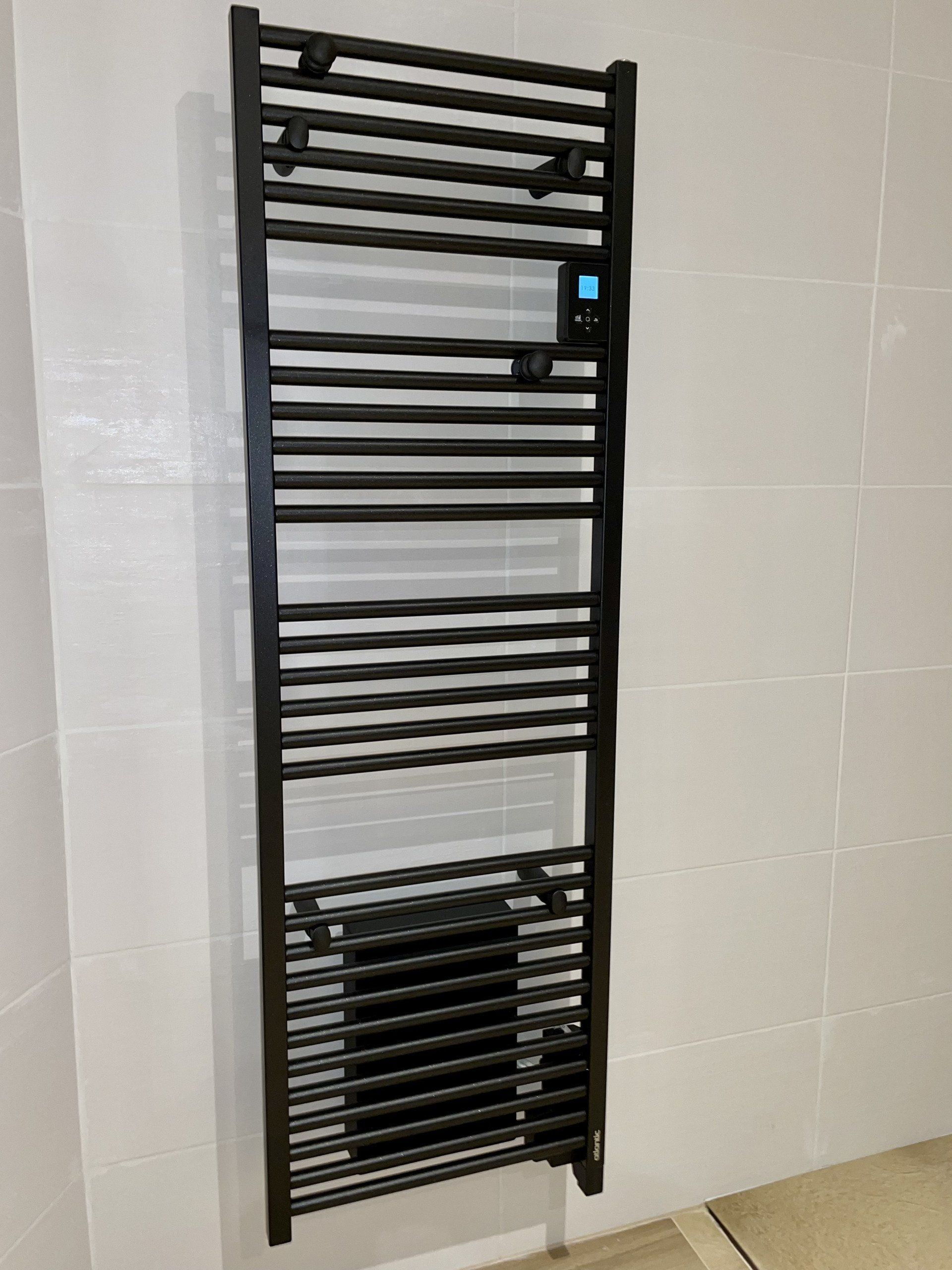 Sèche-serviettes noir fixé sur un mur carrelé blanc. Il est composé de barres horizontales et d'un affichage numérique.