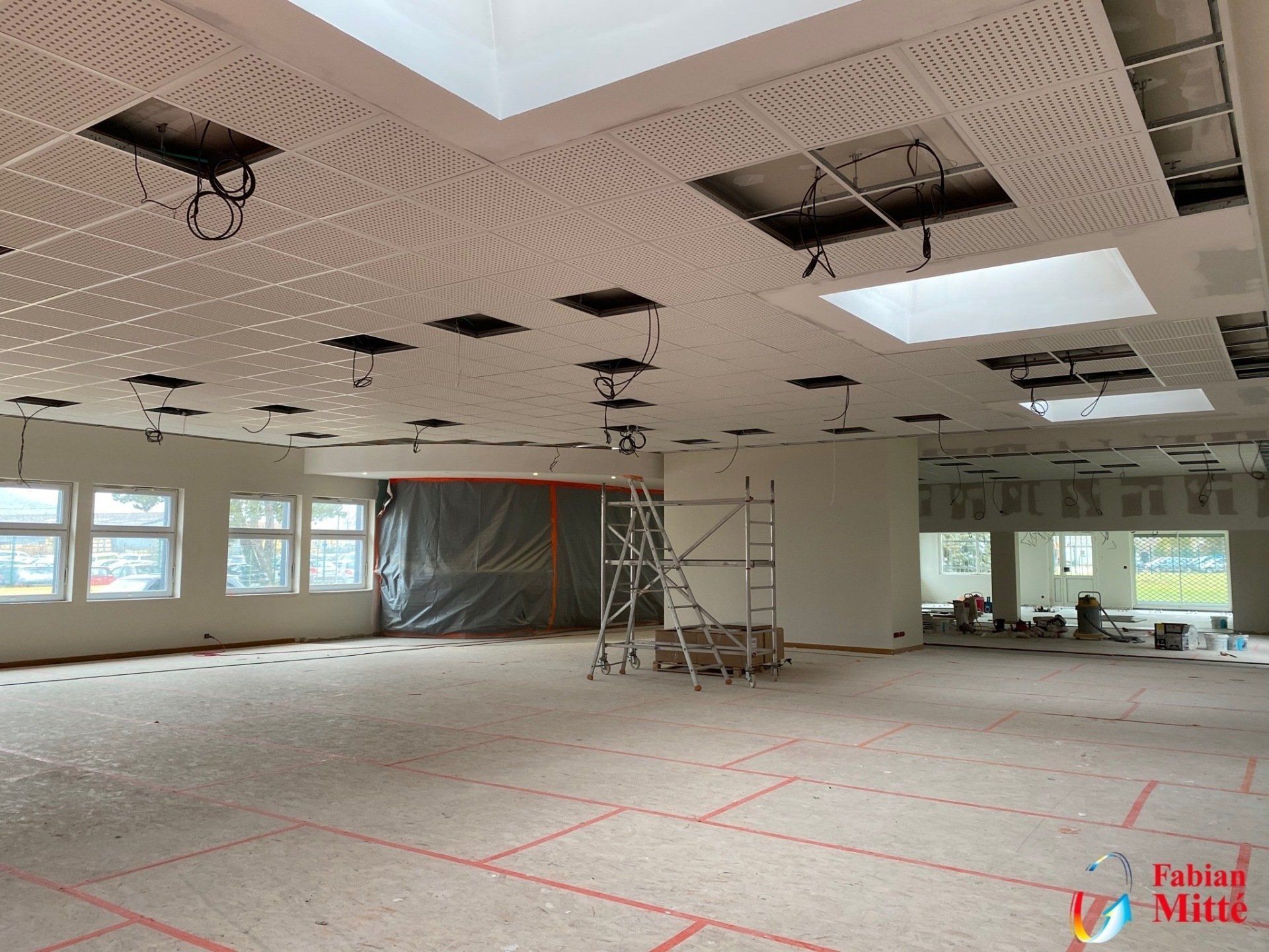 Intérieur d'une pièce en construction avec des panneaux de plafond apparents et un sol non fini. Un échafaudage est visible au centre.
