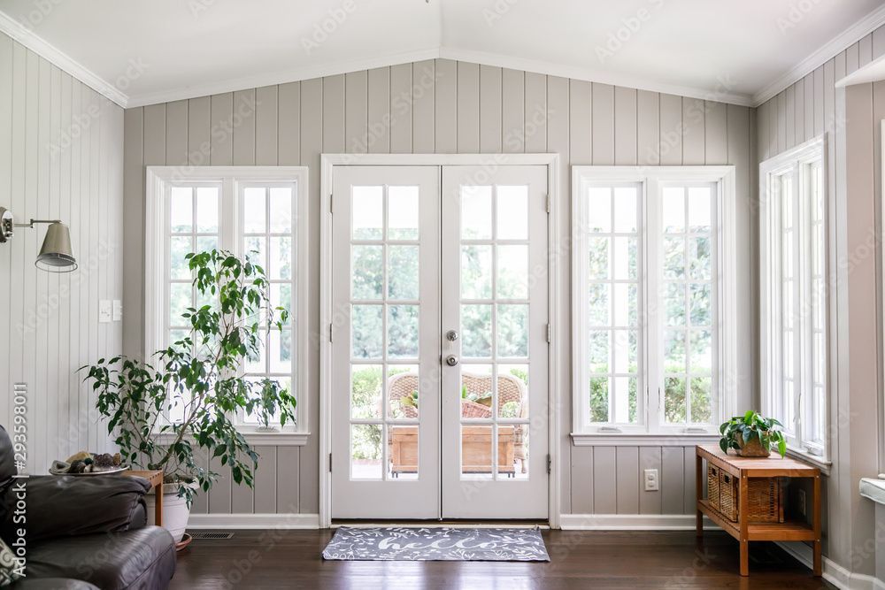 Wintergarten mit weiss gerahmten Fenstertüren und -türen, grau getäfelten Wänden, dunklem Holzboden und Topfpflanzen.