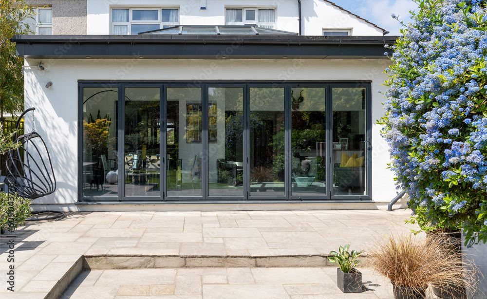 Die Rückseite eines weissen Hauses mit grossen Falttüren aus Glas, die zu einer Terrasse mit Steinstufen und einem blühenden Busch führen.