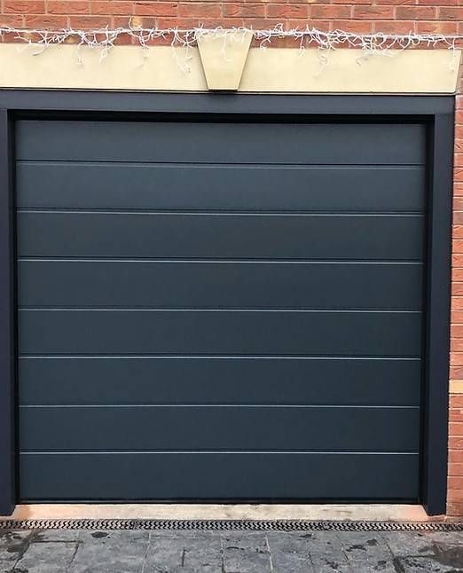 Dunkelgraues, horizontal gepaneeltes Garagentor mit schwarzem Rahmen vor einer Backsteinmauer.