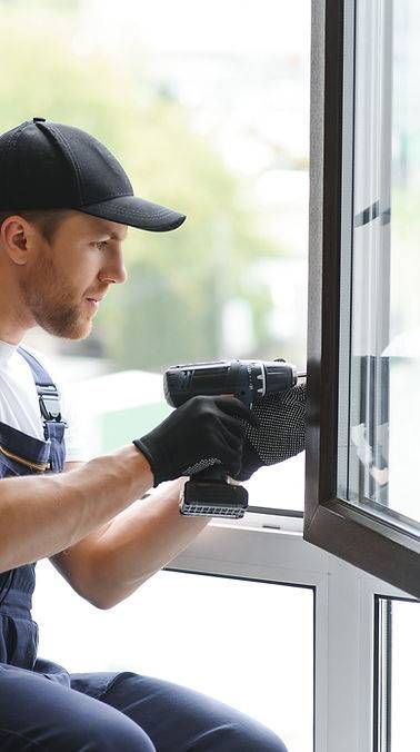 Ein Mann in Latzhose montiert mit einer Bohrmaschine ein Fenster. Er trägt Handschuhe und eine Baseballkappe.