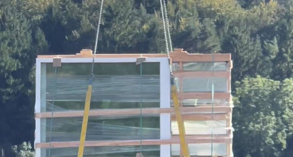 Ein Gebäudemodul mit Glasfenstern und Holzrahmen wird mit einem Kran angehoben.