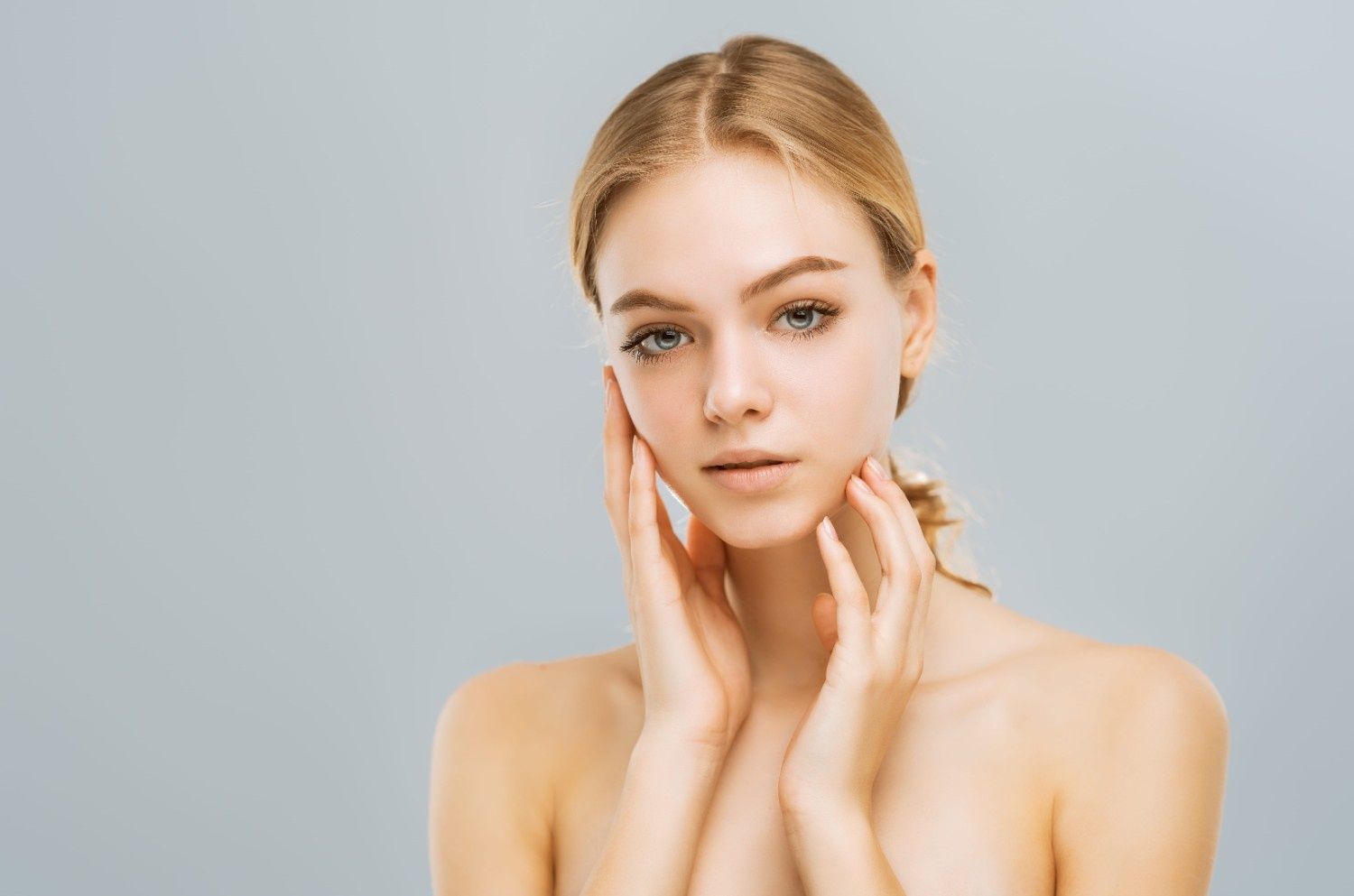 Una persona de cabello rubio y piel tersa se lleva las manos a las mejillas sobre un fondo liso de color gris claro.