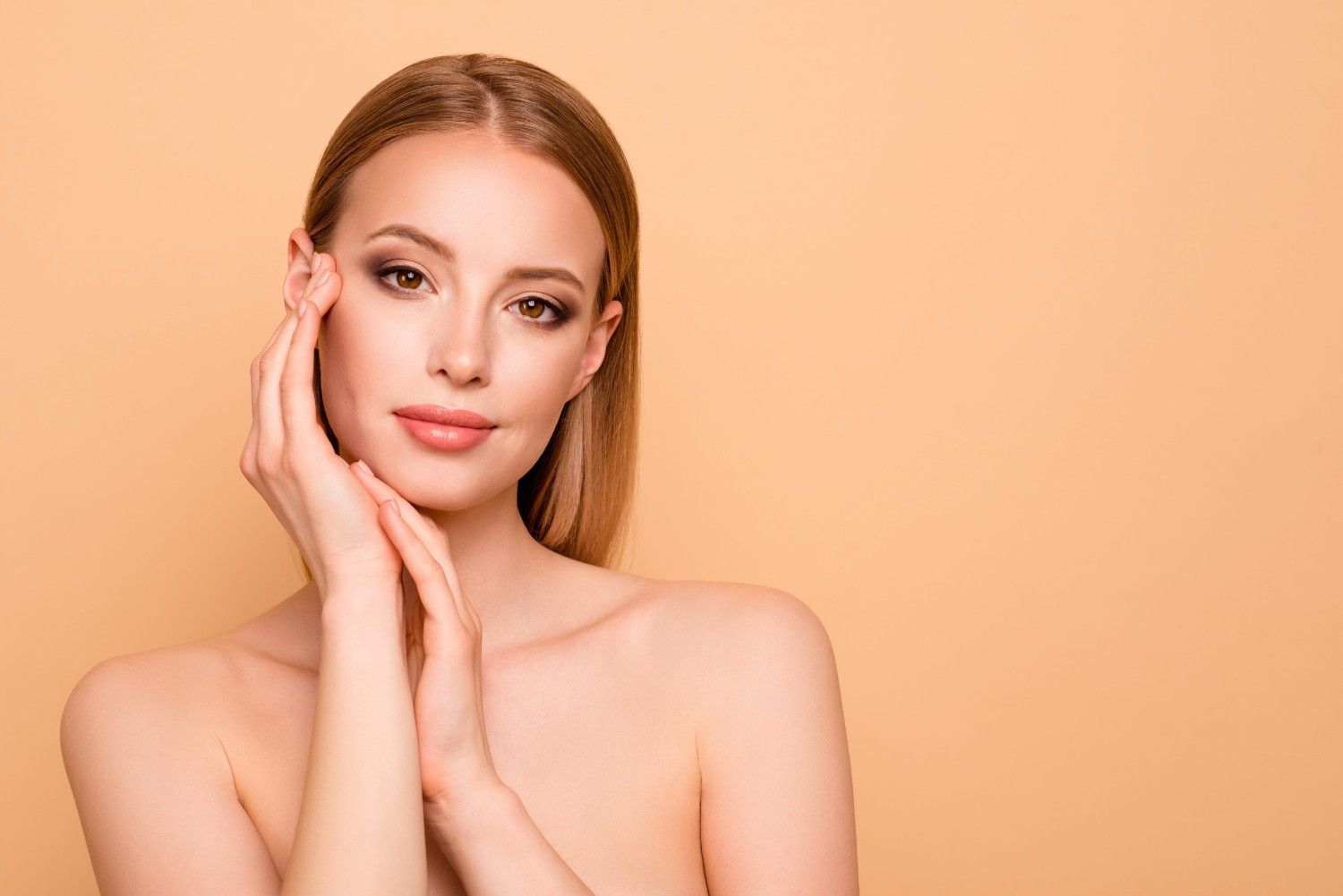 Una persona de tez clara y cabello rubio se cubre el rostro con ambas manos sobre un fondo beige liso.