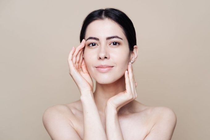 Una persona de piel tersa se toca la cara con ambas manos, sobre un fondo neutro de color beige claro.