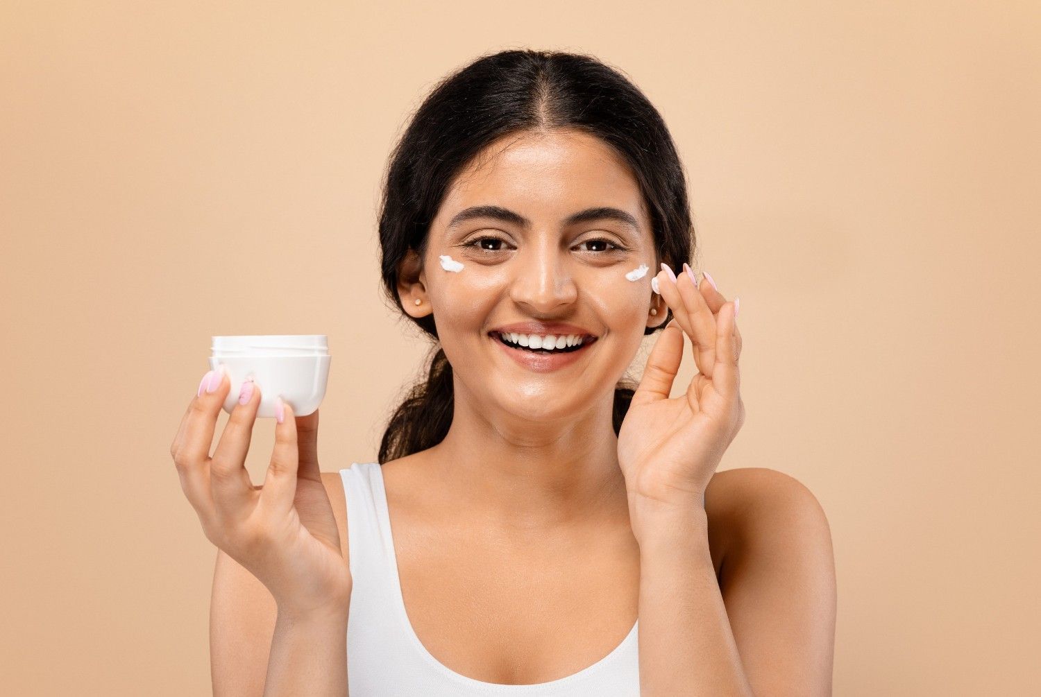 Una persona sonriente se aplica crema facial blanca en la mejilla sobre un fondo beige.