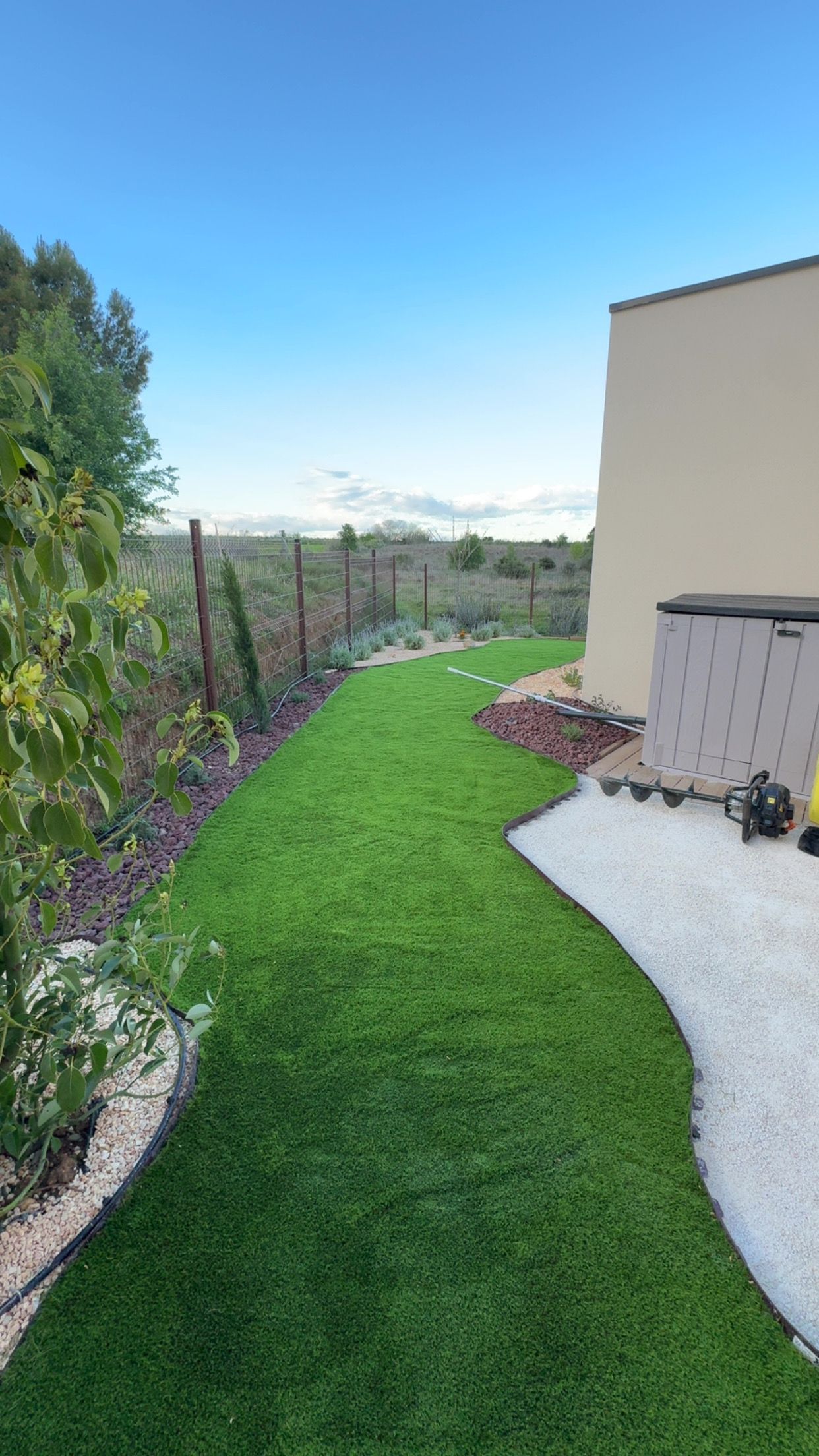 Une bande incurvée de gazon artificiel vert borde un mur beige, du gravier blanc et un paysage naturel sous un ciel bleu.