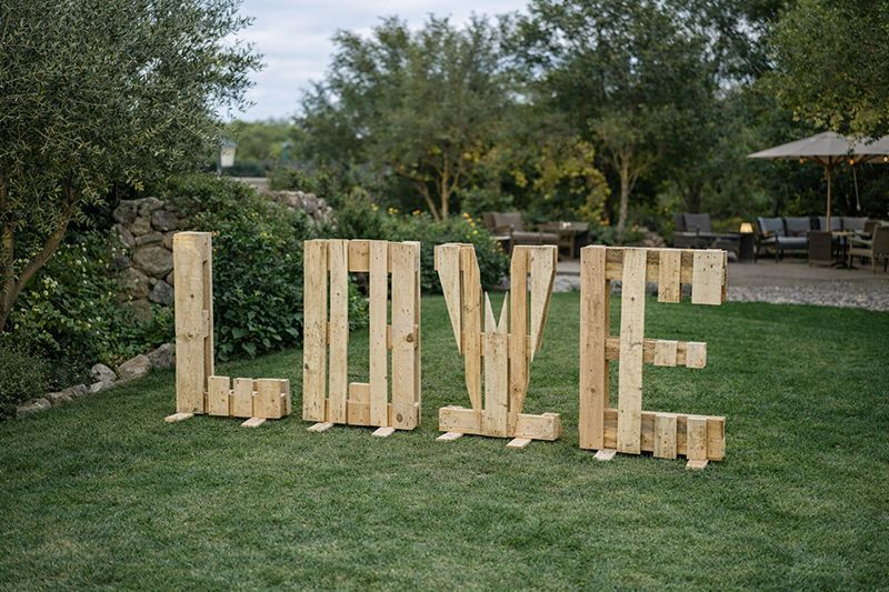 Große, aus Paletten gefertigte Holzbuchstaben, die das Wort „LOVE“ bilden, stehen auf einem grünen Rasen in einem angelegten Garten.