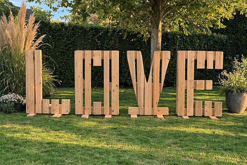 Große Holzbuchstaben, die das Wort „LIEBE“ bilden, stehen auf einer Rasenfläche vor einem Baum und einer Hecke.