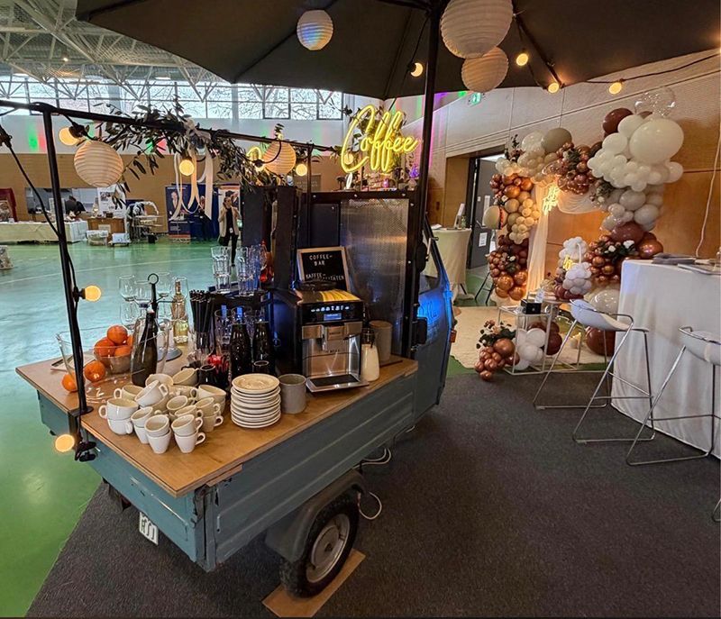 Ein türkisfarbener Kaffeewagen mit Leuchtreklame, Tassen und Orangen steht in einem Veranstaltungsraum in der Nähe, daneben befindet sich ein Ballonbogen.