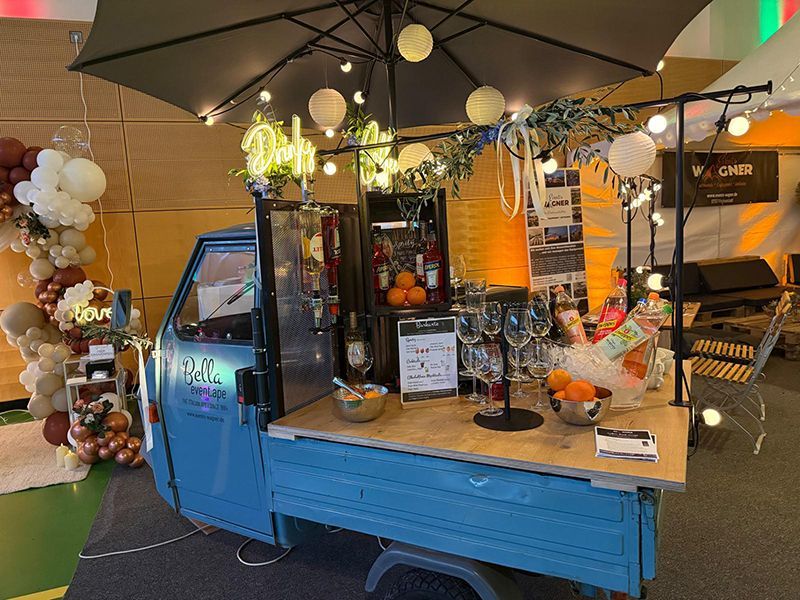 Ein blauer, dreirädriger Oldtimer-Lkw, der zu einer mobilen Bar umgebaut wurde, mit einem großen schwarzen Sonnenschirm, Lichtern und Gläsern.