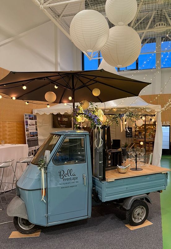 Ein hellblaues, dreirädriges Oldtimerfahrzeug aus Italien, das zu einer Bar umgebaut wurde, steht in einem Innenraum unter einem großen Sonnenschirm und Laternen.