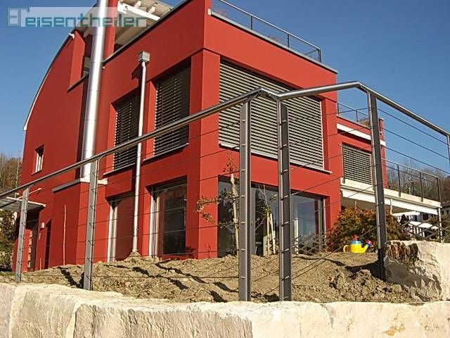 Rotes, modernes Haus mit Edelstahlgeländer und Fensterläden vor blauem Himmel.