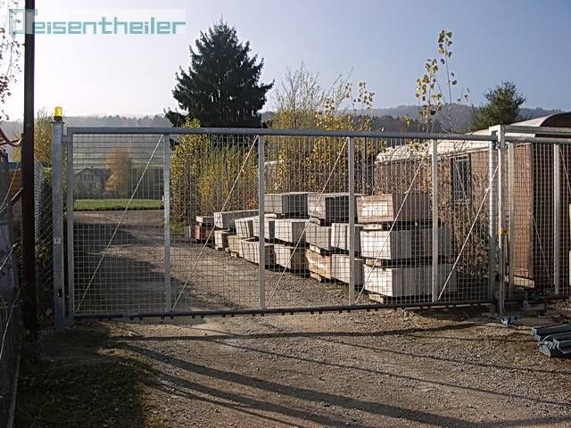 Offenes Sicherheitstor aus Metall mit Maschendrahtgittern umschliesst gestapelte Betonblöcke. Freiluftkulisse mit Bäumen und Himmel.