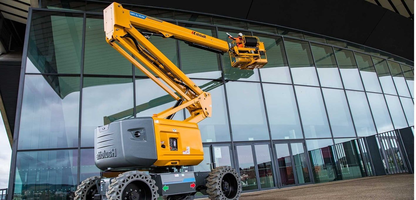 Plataforma elevadora amarilla que sube a un edificio con paredes de cristal; hay una persona en la plataforma.