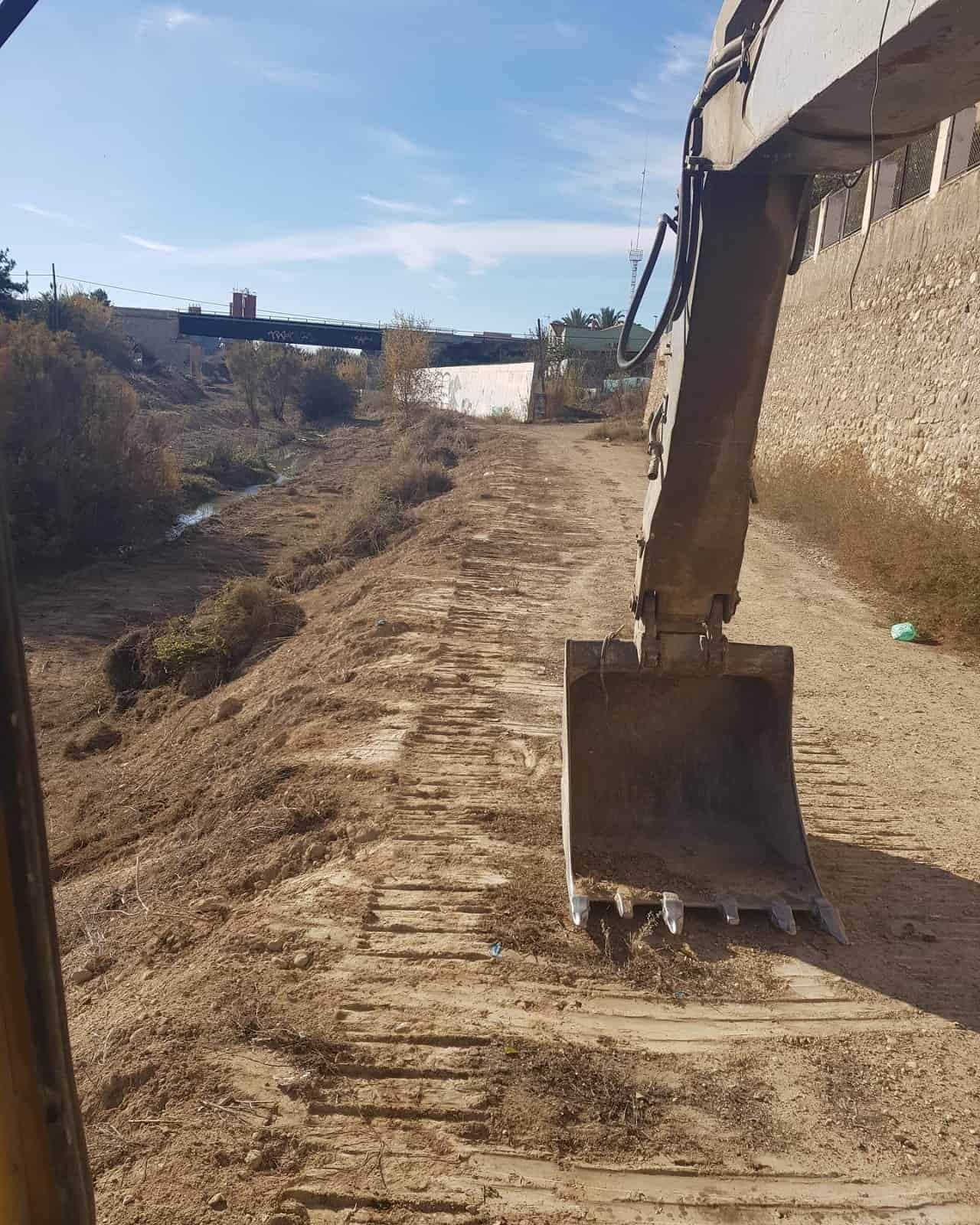 Brazo de excavadora en una zanja de tierra marrón, despejando el camino. Soleado, con un terraplén alto.