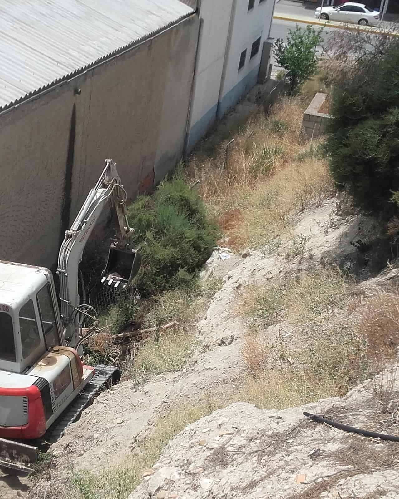 Una excavadora estacionada en una zanja seca y cubierta de vegetación al lado de un edificio.