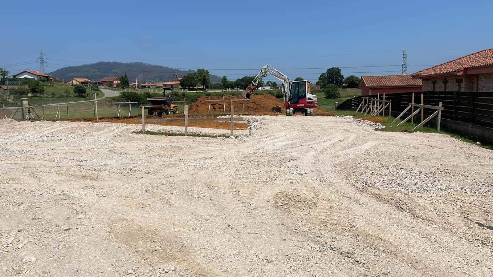 Sitio de construcción con excavadora y montón de tierra, grava en primer plano, edificios y una colina.