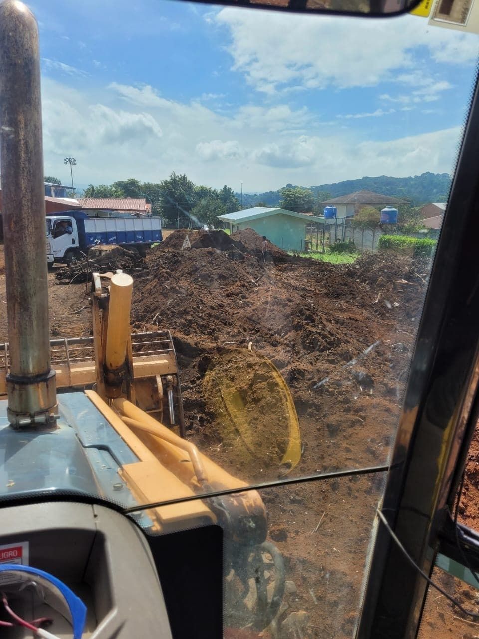 Vista desde el interior de la cabina de una excavadora, mirando una pila de tierra, edificios y un cielo parcialmente nublado.