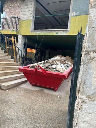 Cargadora amarilla trabajando en un camino de tierra. Una persona está al volante. Edificio al fondo.