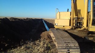 Excavadora cavando una zanja larga y recta en un campo bajo un cielo azul.