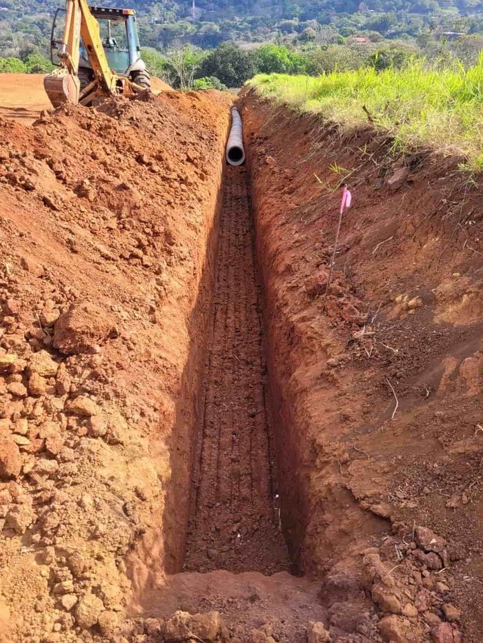 Zanja cavada en tierra roja con una tubería instalada; excavadora al fondo, césped y colinas más allá.