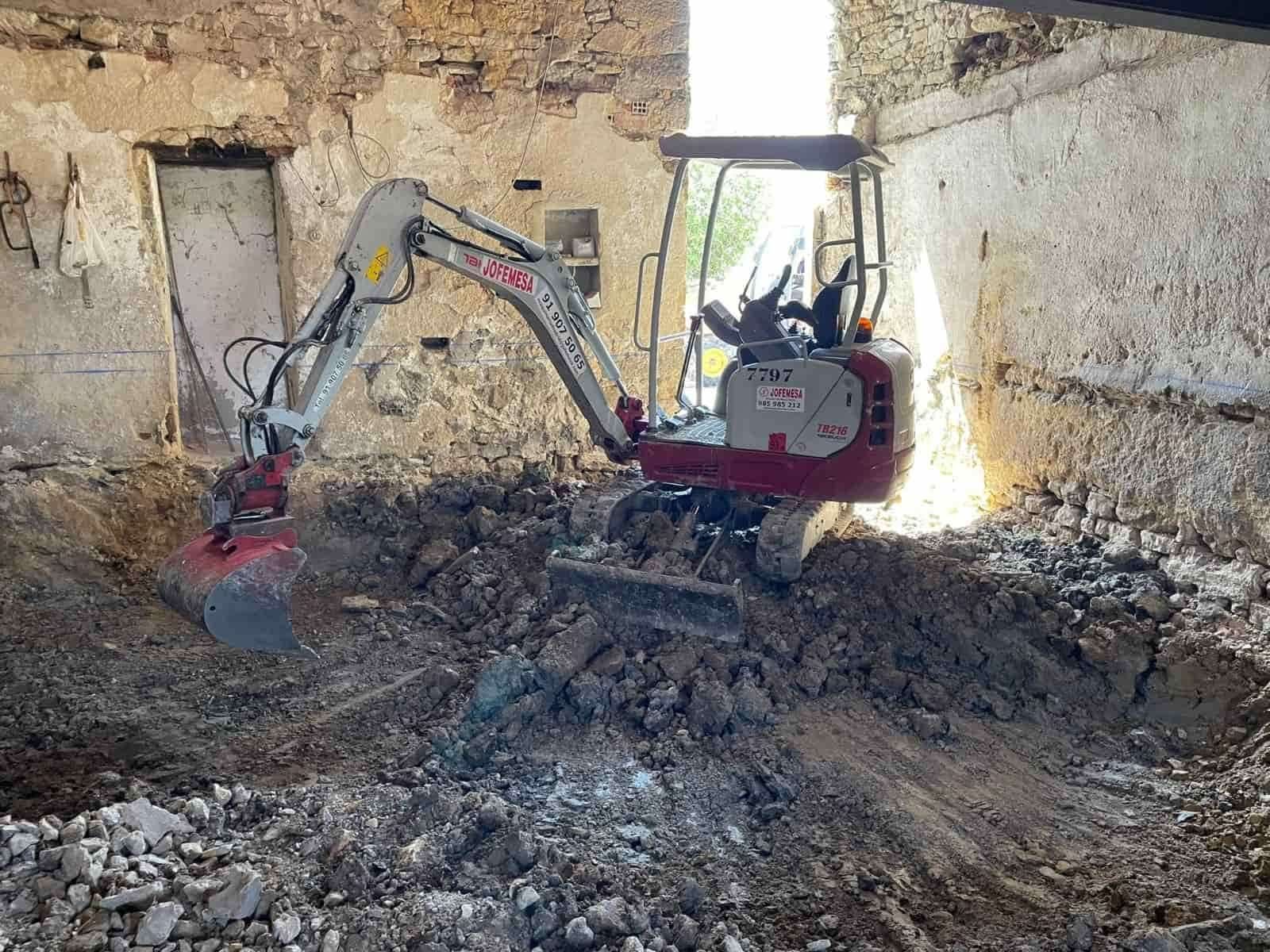 Miniexcavadora cavando tierra dentro de un edificio con paredes de piedra y una puerta.